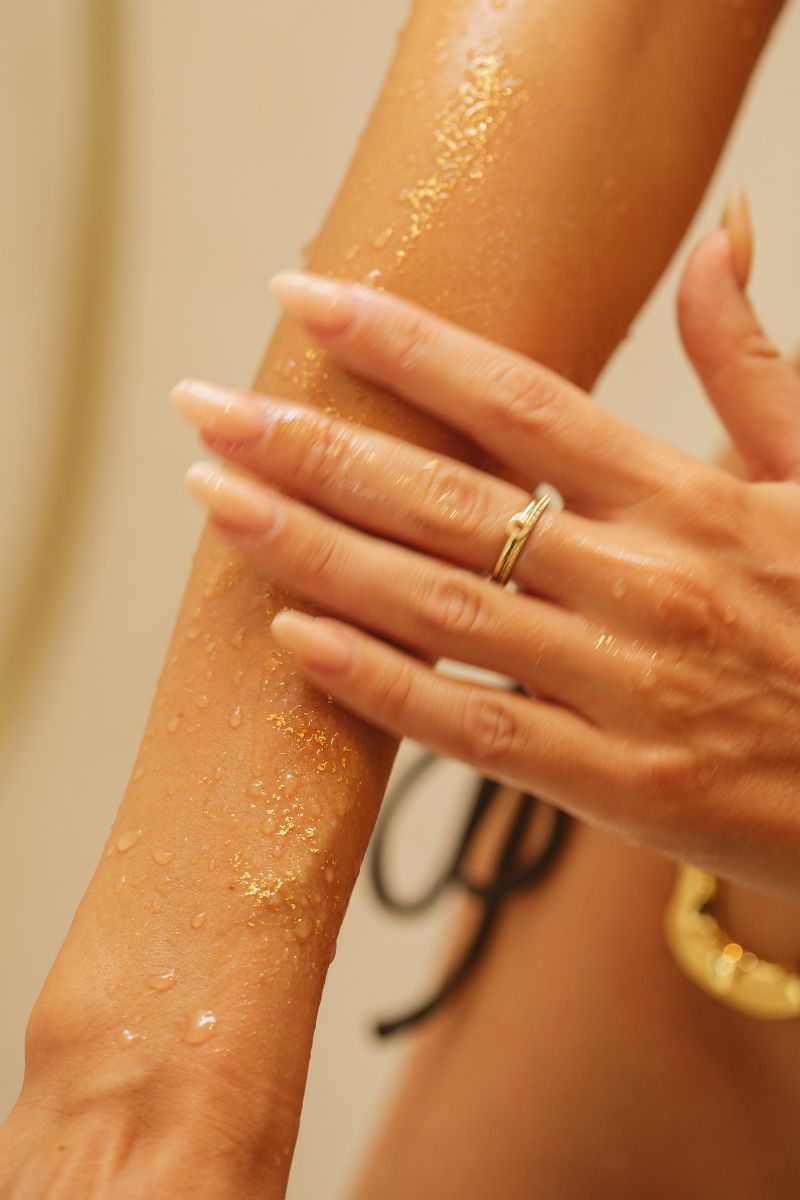 Woman applying UMM Skincare's lavish bath products, the body oil, to her arm with gold flakes making her skin sparkle.
