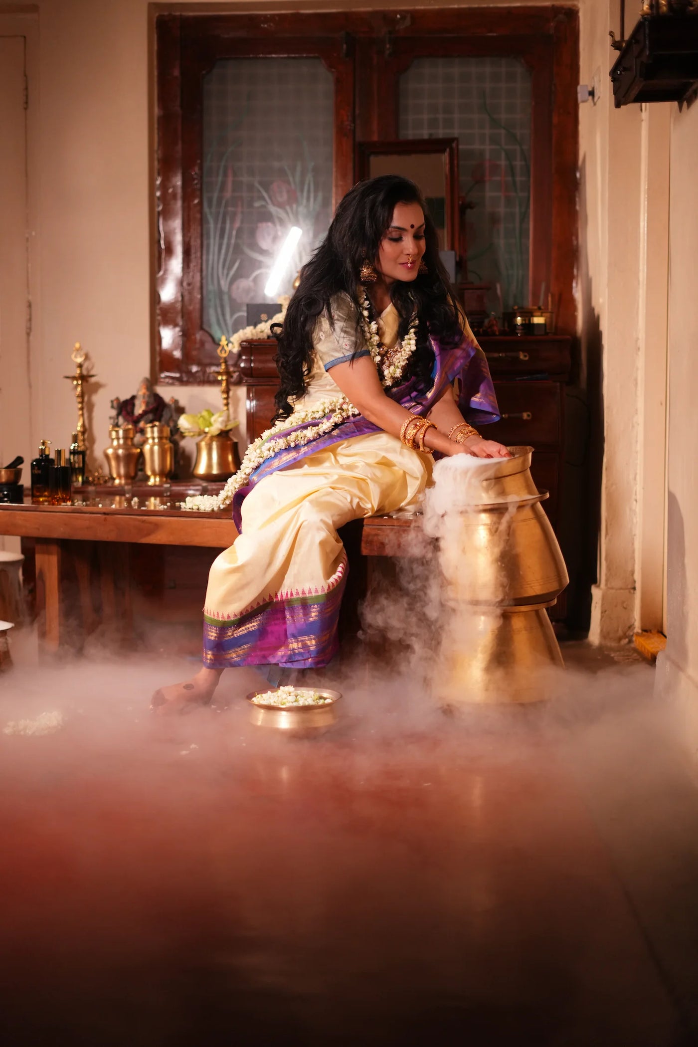 Anisha seated beside a brass vessel, in ritual dress with a floral garland, steam rising around her
