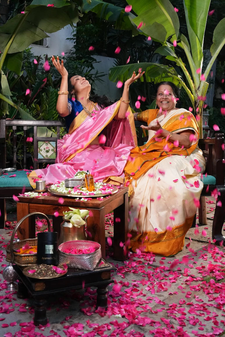 Anisha with an older family member, laughing beneath banana leaves as flower petals fall around them